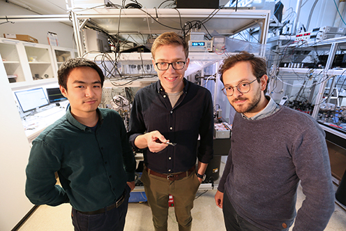 A thin silicon nitride membrane (white) is stretched tight across a silicon frame (blue). The membrane contains a pattern of holes, with one small island in the center, whose vibrations are measured in the experiment. PhD students Junxin Chen and Massimiliano Rossi on either side of David Mason, first author on the article in Nature Physics. David is holding the silicon nitride membrane in his hand in a pair of tweezers. Junxin Che, Massimiliano Rossi and David Mason